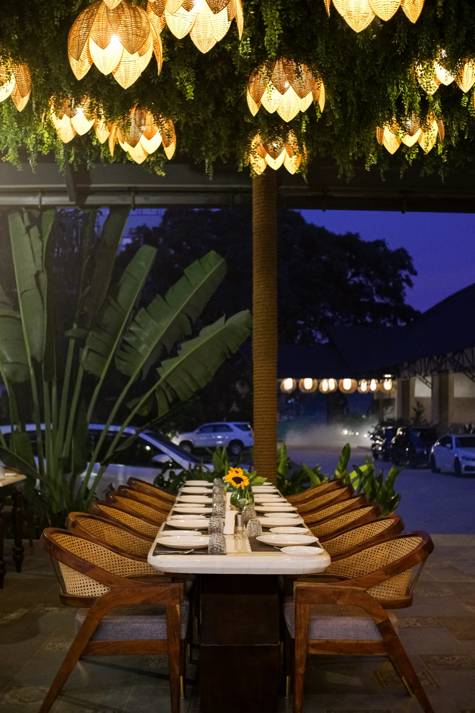 A long restaurant table set for dinner beneath woven pendant lamps at night